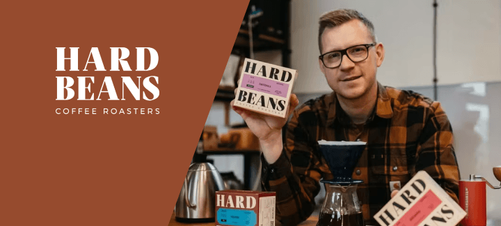 Grafika promocyjna marki Hard Beans podzielona ukośną linią na dwie sekcje. Lewa część to gładkie, rdzawobrązowe tło z białym logotypem tekstowym: duże słowa "HARD BEANS" zapisane szeryfowym fontem, a pod nimi mniejszy napis "COFFEE ROASTERS". Prawa część to fotografia przedstawiająca mężczyznę w czarnych, prostokątnych okularach i brązowo-czarnej koszuli w kratę. Mężczyzna patrzy w obiektyw i trzyma w prawej dłoni kwadratowe, jasne opakowanie kawy Hard Beans z fioletową etykietą informacyjną. Na drewnianym blacie przed nim znajduje się zestaw do alternatywnego parzenia kawy: szklany serwer z nałożonym ciemnoniebieskim dripperem, czerwony młynek ręczny oraz dwa dodatkowe kartoniki z kawą marki Hard Beans (jeden z błękitną, a drugi z różową etykietą). W tle widoczny jest stalowy czajnik z gęsią szyją oraz rozmyte półki z wyposażeniem.