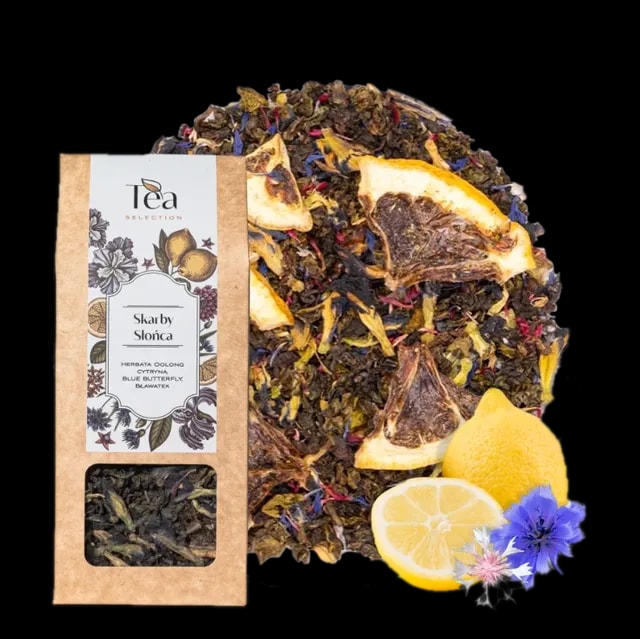 Opakowanie herbaty Skarby Słońca na białym tle, zawierającej mieszankę herbaty oolong, cytryny, blue butterfly oraz bławatera, obok talerz z suszonymi owocami i kwiatami, połowa cytryny, całe cytryny oraz niebieskie kwiaty chabra i białe kwiaty bławatera