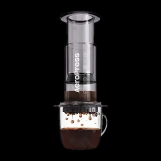 Zdjęcie czarnego zaparzacza AeroPress Clear Black, umieszczonego na szklance z zaparzaną kawą. Krople kawy spadają do kubka, ukazując proces parzenia metodą AeroPress, z przezroczystym czarnym korpusem urządzenia.