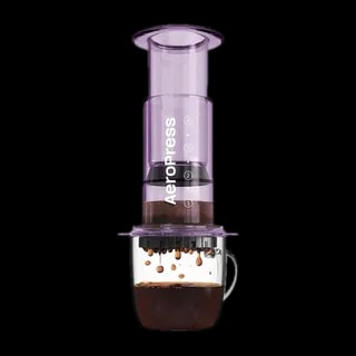 Zdjęcie fioletowego zaparzacza AeroPress Clear Purple, umieszczonego na szklance z zaparzaną kawą. Krople kawy spadają do kubka, ukazując proces parzenia metodą AeroPress, z przezroczystym fioletowym korpusem urządzenia.