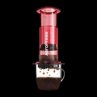 Zdjęcie czerwonego zaparzacza AeroPress Clear Red, umieszczonego na szklance z zaparzaną kawą. Krople kawy spadają do kubka, pokazując proces parzenia metodą AeroPress, z przezroczystym czerwonym korpusem urządzenia.