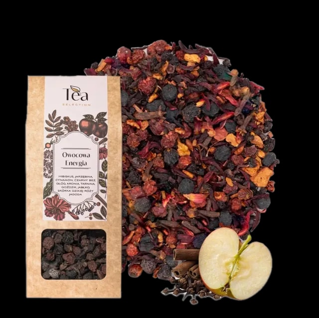 Opakowanie herbaty Owocowa Energia na białym tle, zawierającej mieszankę hibiskusa, jarzębiny, cynamonu, czarnego bzu, głogu, aronii, tarniny, goździków, jabłka, skórki dzikiej róży i jałowca, obok talerz z suszonymi owocami i przyprawami, pół jabłka, laska cynamonu oraz goździki